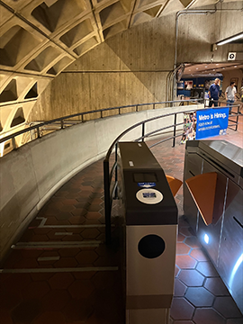 A picture of a pathway. To the right in the foreground is a faregate and to the left is the pathway. Along the pathway, which curves to the right in the background, there is a railing on the right hand side and a side wall with a railing on the left hand side.